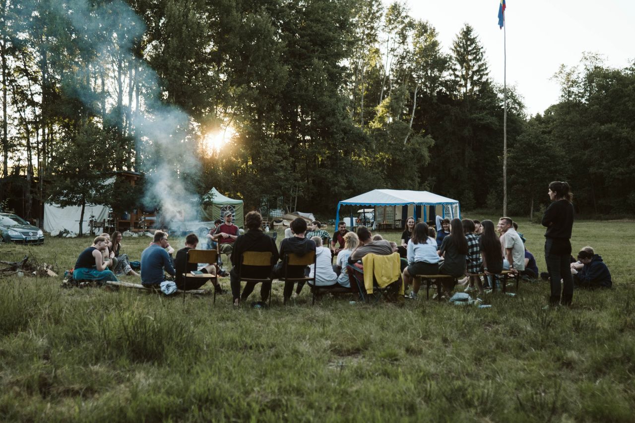 A group of adults relaxing around a campfire in a scenic outdoor setting with tents.